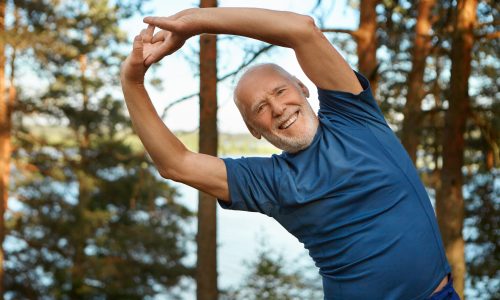 Outdoor shot of happy energetic senior retired man enjoying physical training in park, doing side bends exercise, holding hands together, looking at camera with broad smile, warming up body before run